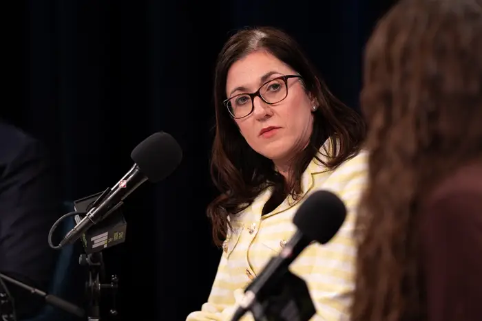 Jessica Katz, the city's chief housing officer, sits in front of a microphone while wearing a yellow and white striped blazer.